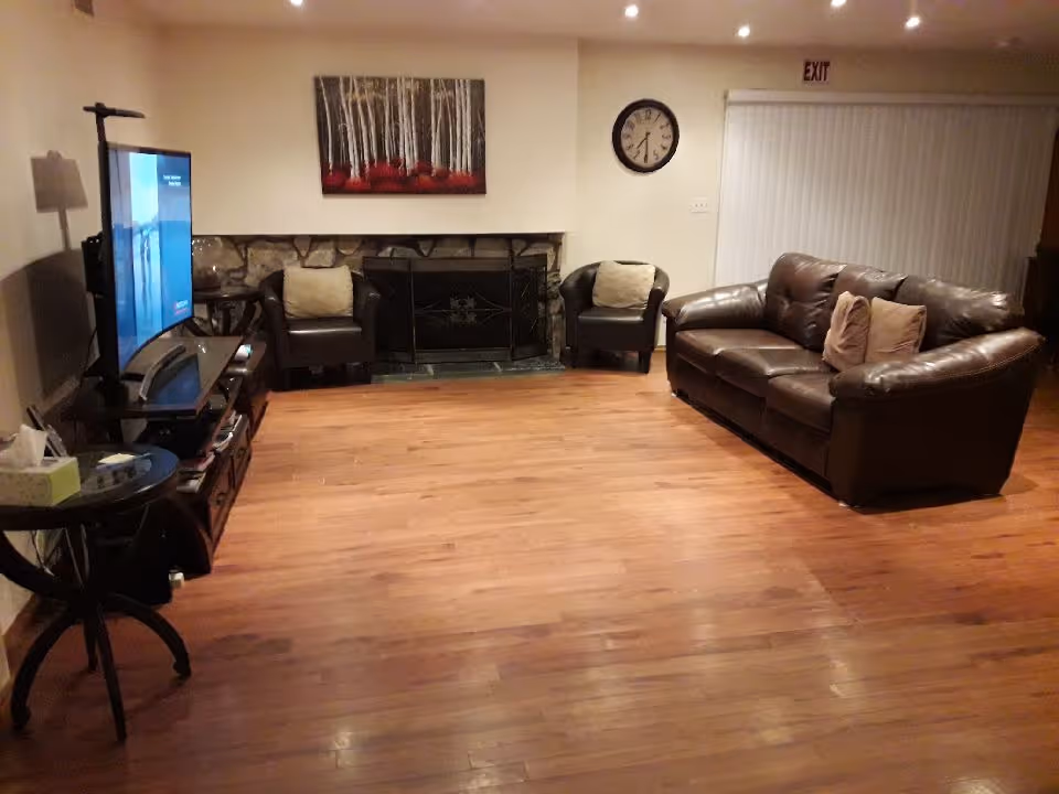 A cozy living room with wooden flooring, a brown leather sofa with two cushions, two black armchairs with beige cushions, a stone fireplace with a black metal screen, a wall clock, a large flat-screen TV on a stand, a side table with a tissue box, and a painting of birch trees with red foliage above the fireplace.