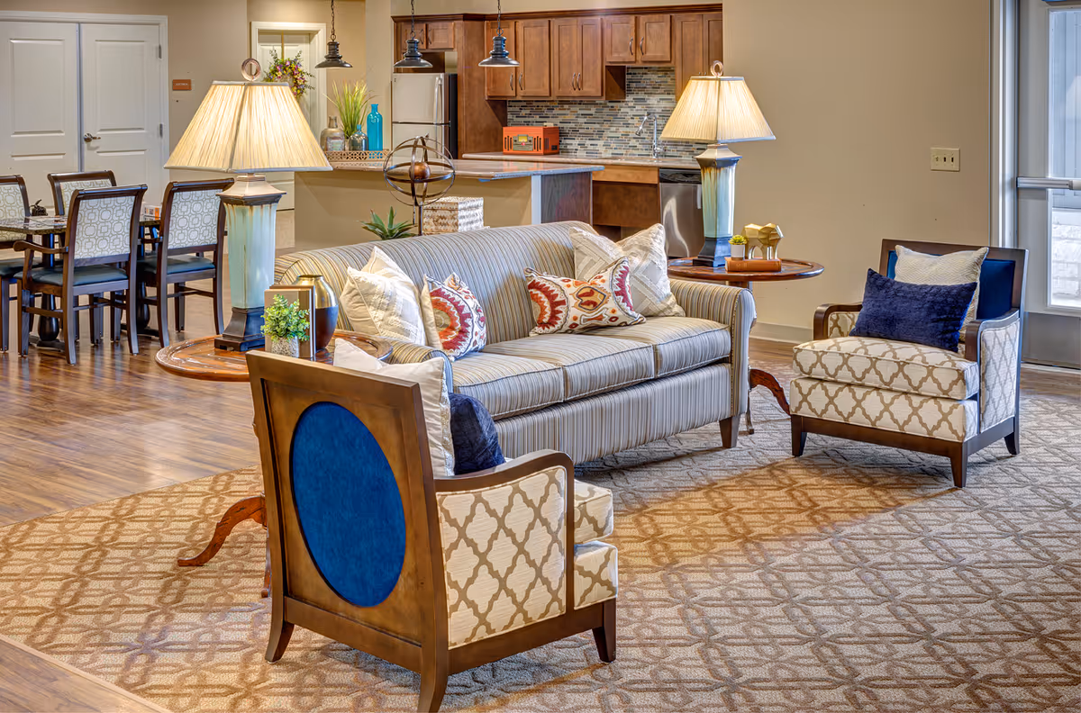 A cozy living room area in The Delaney at Georgetown Village featuring a striped sofa with decorative pillows, two patterned armchairs with blue cushions, two table lamps on side tables, a patterned carpet, and a view into a kitchen with wooden cabinets and stainless steel appliances. A dining area with chairs and a table is visible in the background.