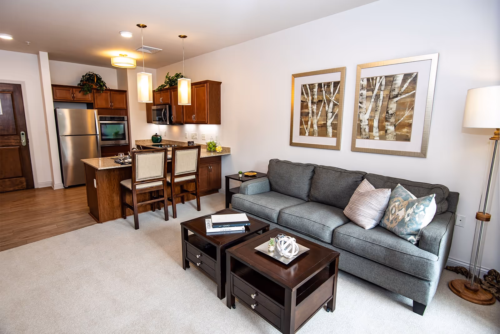 A modern living room and kitchen area in a senior living facility. The living room features a gray sofa with three decorative pillows, two framed pictures of birch trees on the wall, a floor lamp, and two dark wooden coffee tables with decorative items. The kitchen has wooden cabinets, stainless steel appliances including a refrigerator and oven, a small island with two chairs, and pendant lights hanging above the island.