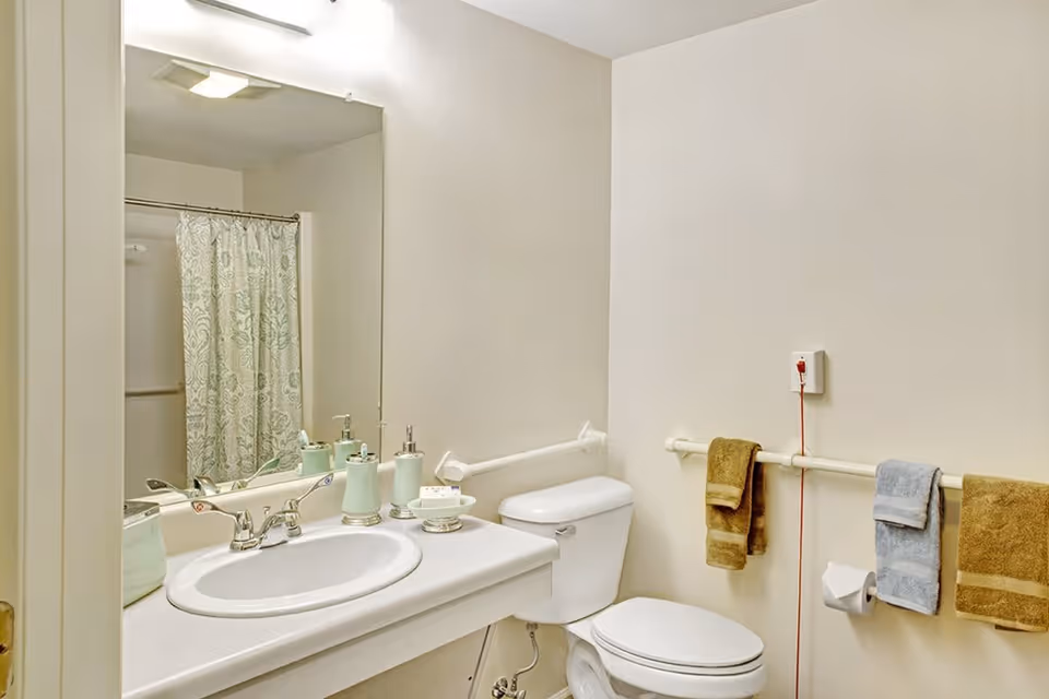 A clean bathroom with a white sink and countertop, a large mirror above the sink, a toilet, and a shower with a patterned curtain. There are three towels hanging on a grab bar next to the toilet, and a red emergency pull cord is visible on the wall.