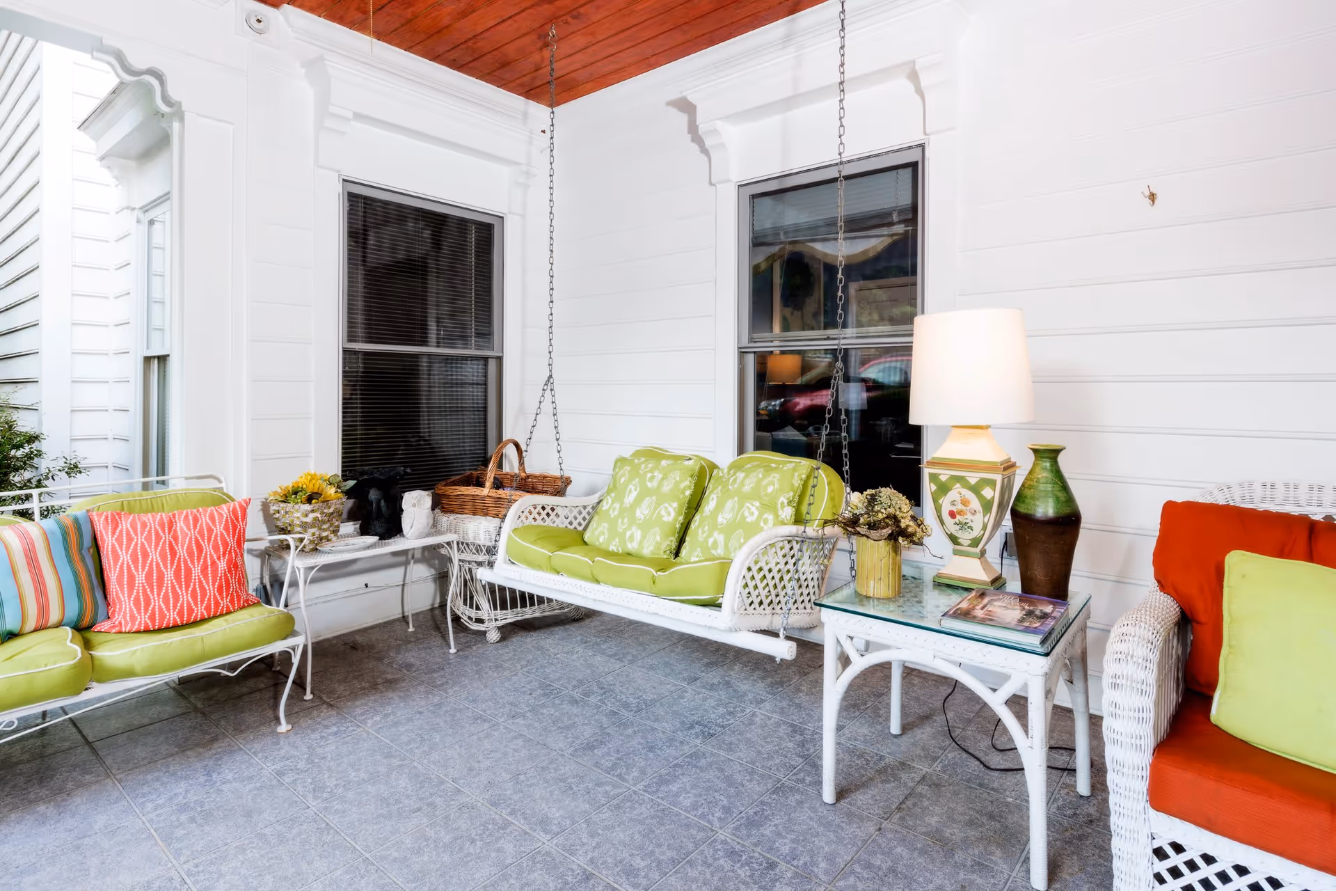 A cozy porch area with white walls and a wooden ceiling. The porch features a white metal bench with green cushions and colorful pillows, a white hanging swing with green cushions, a white wicker chair with orange and green cushions, and a white side table with a lamp, a green vase, and a small plant. There are two windows with black blinds and decorative items including a basket and a small statue on a white table.
