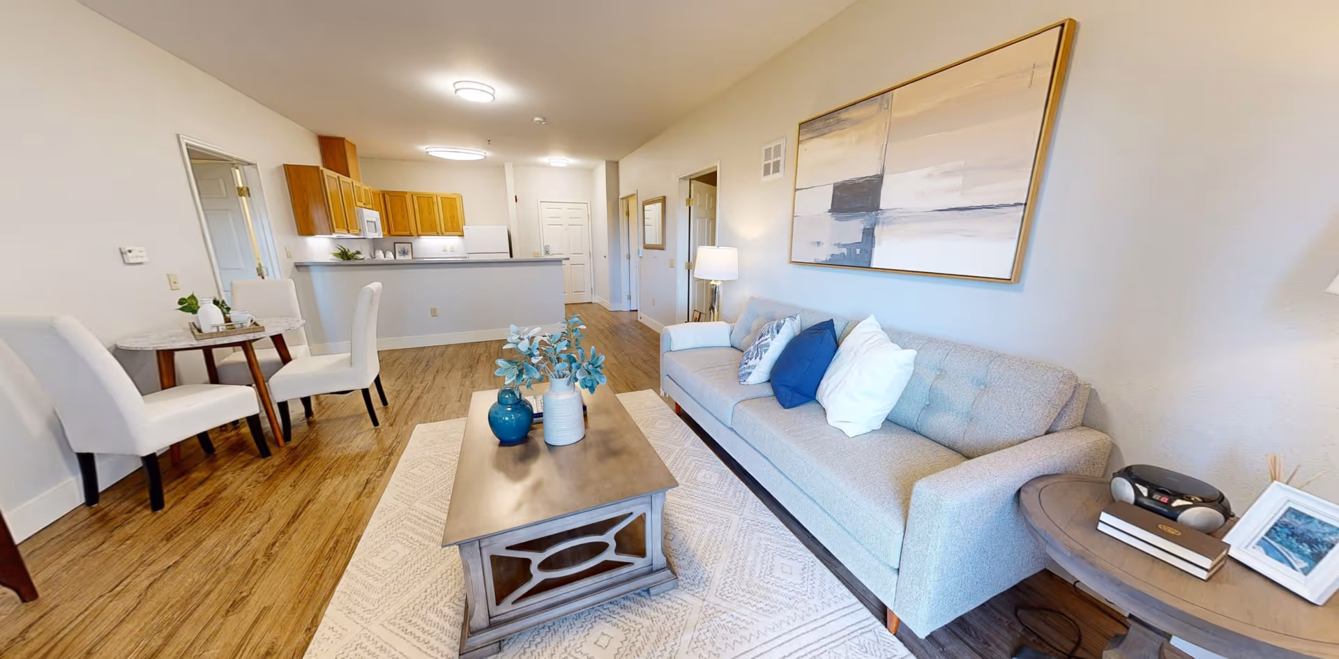 A bright living room with a light gray sofa adorned with white and blue pillows, a wooden coffee table with decorative vases, a round side table with books and a small stereo, and a small dining area with two white chairs and a round table. The room has wooden flooring and a kitchen with wooden cabinets and white appliances in the background.