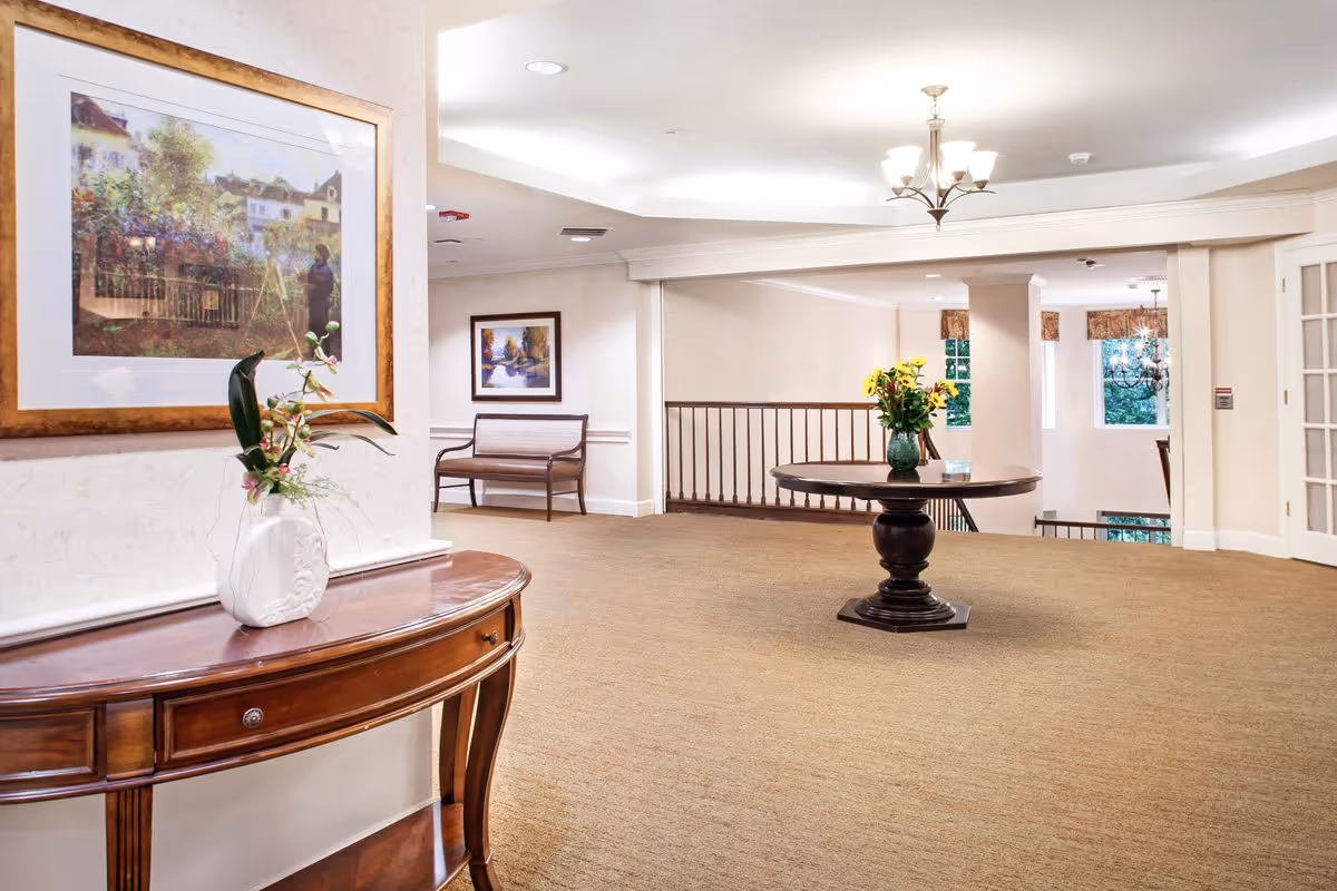 A spacious, well-lit interior hallway area with beige carpet and cream-colored walls. There is a wooden console table with a white vase holding flowers on the left side, and a round wooden table with a green vase of yellow flowers in the center. Two framed paintings hang on the walls, and a wooden bench is placed against the far wall. A chandelier hangs from the ceiling, and windows allow natural light to enter the space.
