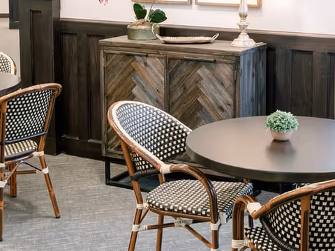 Small dining area with round tables, woven bistro chairs, and a wooden sideboard against a paneled wall.