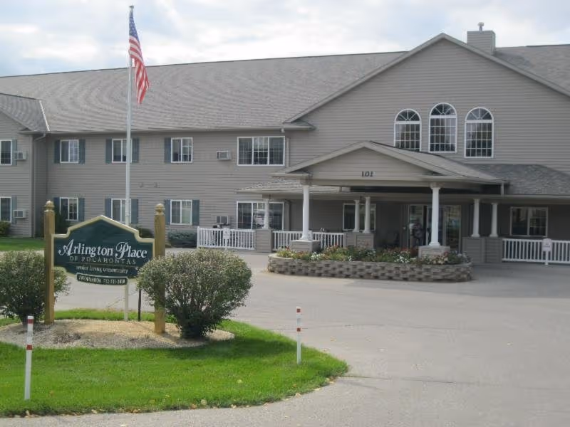 Exterior view of Arlington Place at Pocahontas senior living community building with a green sign in front, an American flag on a flagpole, and a driveway leading to the entrance with a covered porch and flower bed.