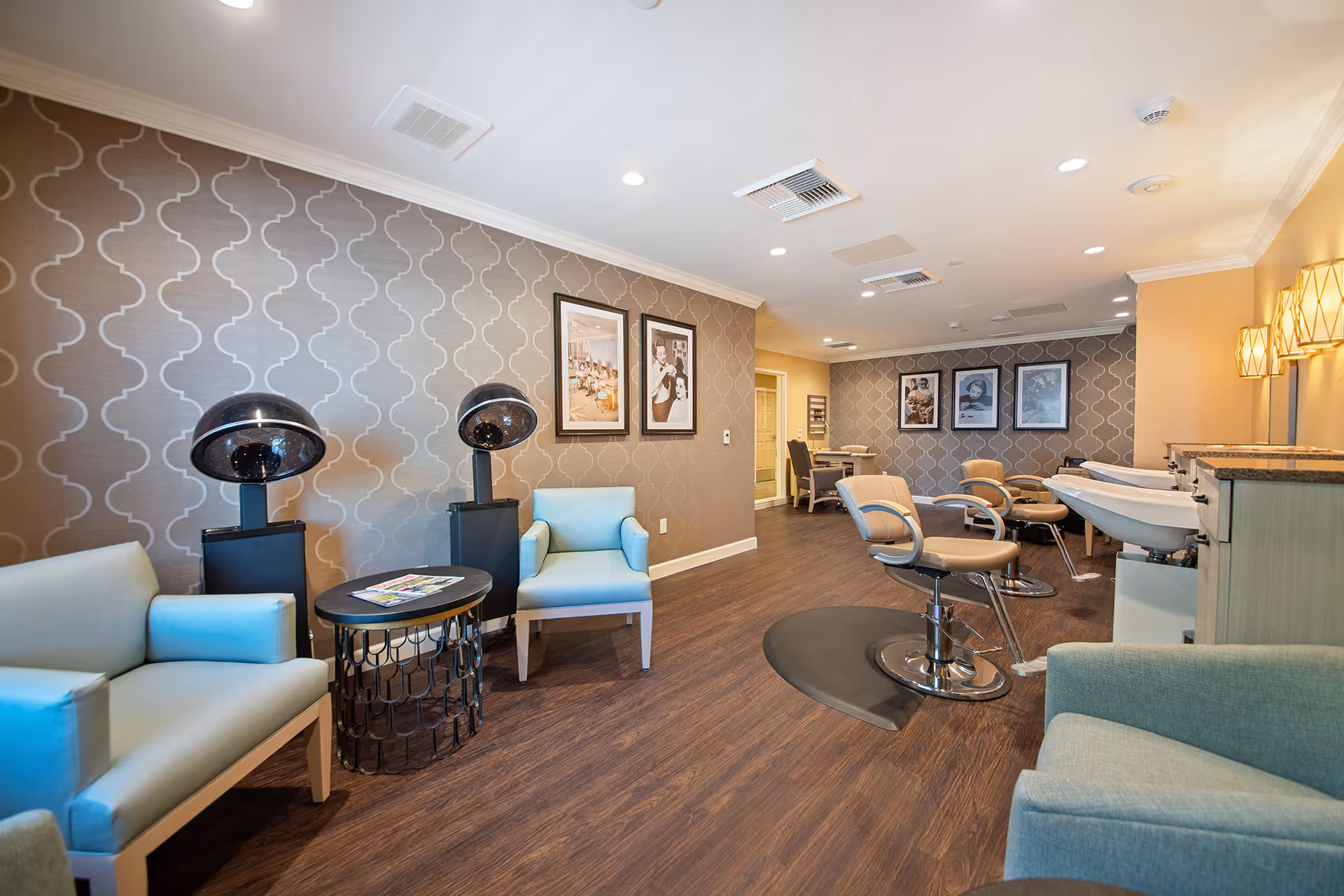 Interior view of a salon area in a senior living facility with light blue armchairs, hair drying stations, salon chairs, sinks for hair washing, and framed pictures on patterned walls. The floor is wooden, and the room is well-lit with ceiling lights and wall sconces.