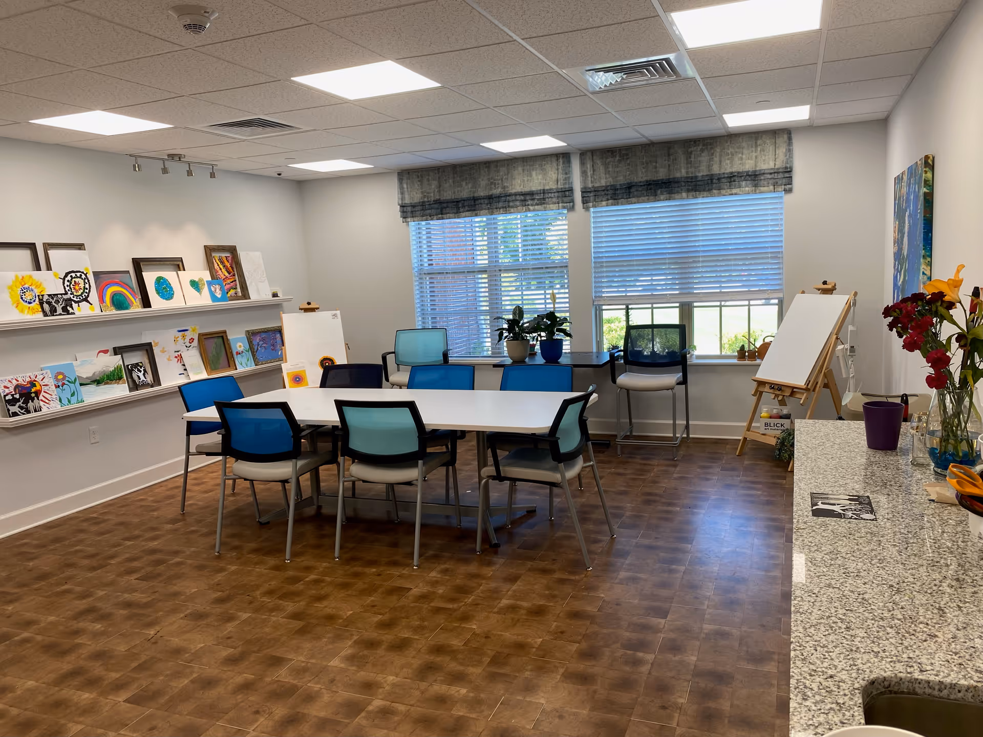 A bright art activity room with a rectangular table surrounded by chairs with blue and teal backs. The walls display various colorful paintings and artwork on shelves. Large windows with blinds allow natural light to fill the room. There is an easel with a blank canvas near the windows and a countertop with flowers and art supplies on the right side.