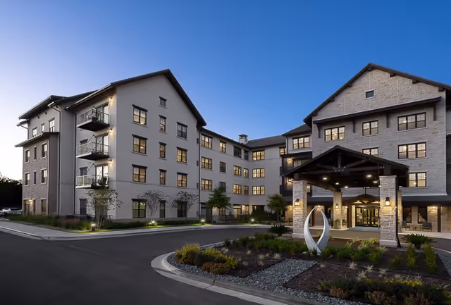 Exterior view of a modern multi-story senior living facility building at dusk with well-lit entrance, landscaped garden, and clear sky.