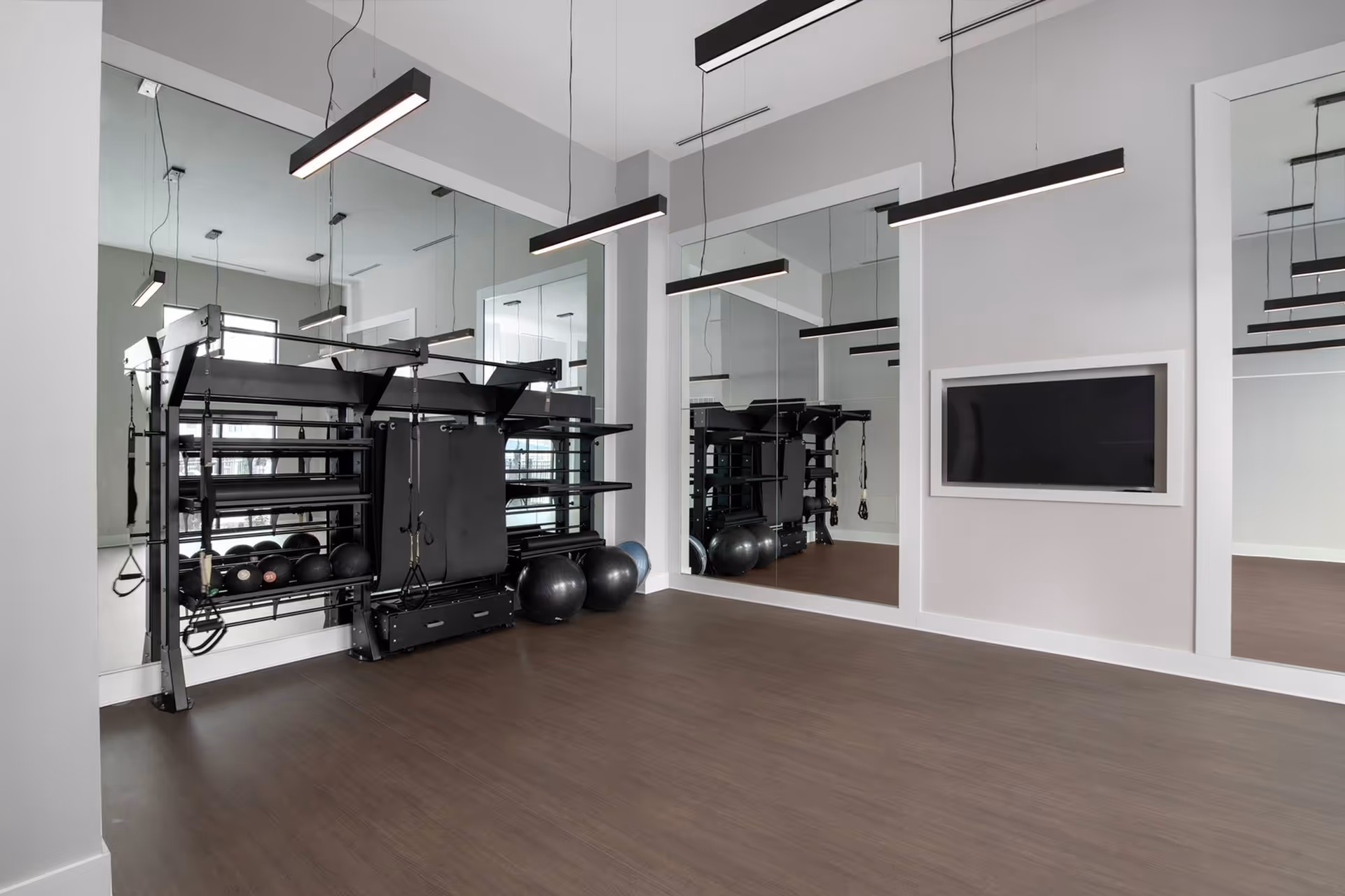 A modern fitness studio with mirrored walls, an equipment storage rack holding medicine balls and accessories, a wall-mounted TV, and suspended linear light fixtures over a wood floor.