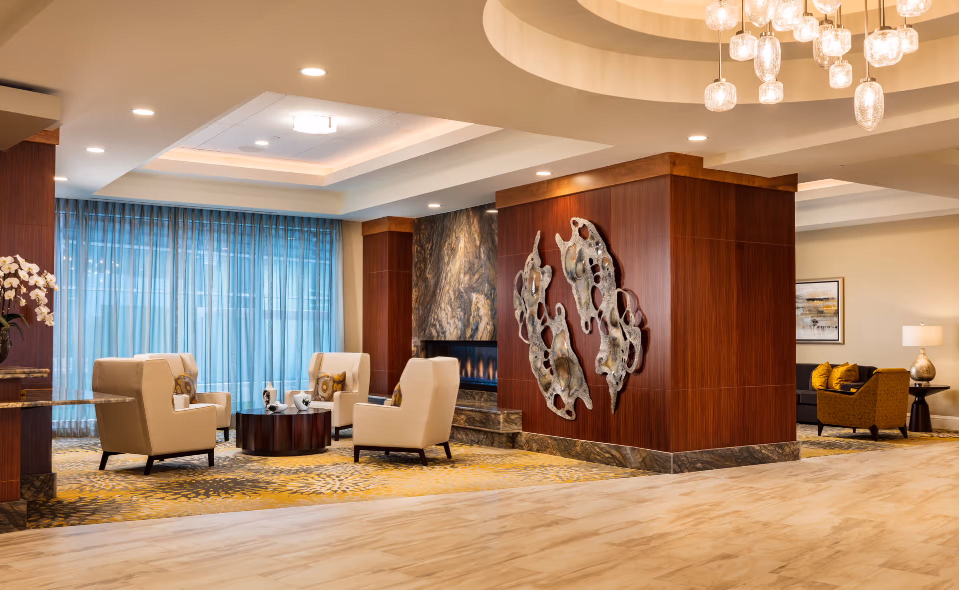 A spacious and elegant living room area in a senior living facility with beige armchairs arranged around a circular wooden coffee table. The room features a modern fireplace with a marble surround, wood-paneled walls, and decorative metal wall art. Large windows with sheer blue curtains allow natural light to fill the space. A chandelier with multiple glass pendant lights hangs from the ceiling, and additional seating with yellow cushions is visible in the background.