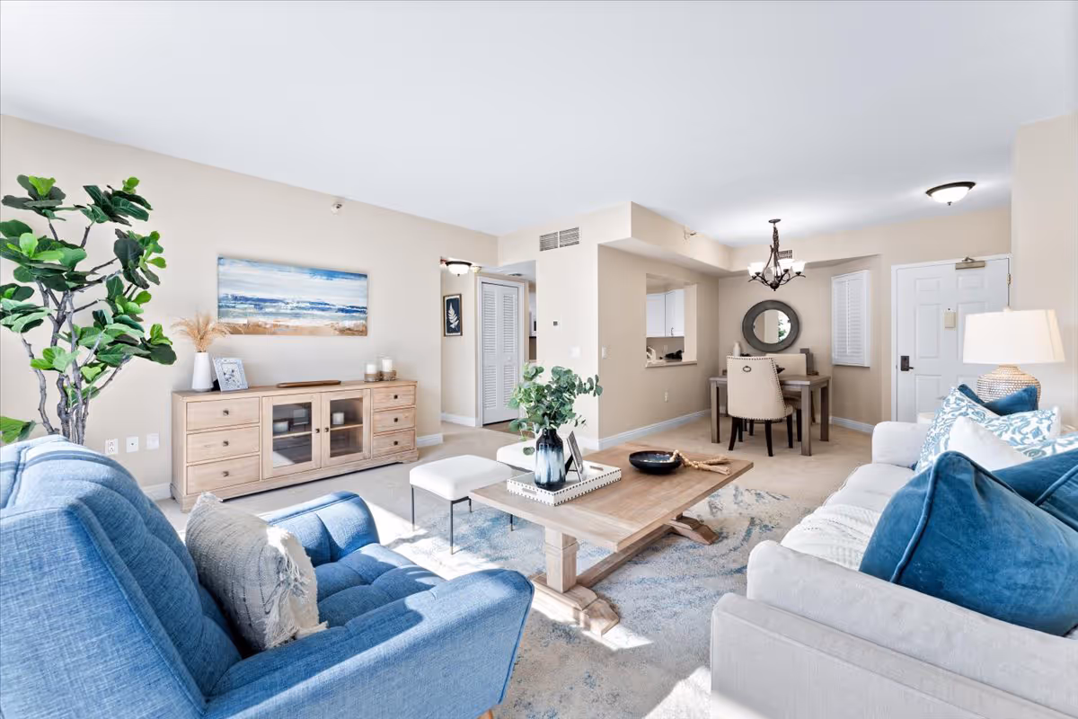 Bright and airy living room and dining area in a senior living facility. The living room features a blue armchair, a beige sofa with blue and white cushions, a wooden coffee table with decorative items, and a wooden sideboard with candles and a framed picture. A large green plant is placed near the sideboard. The dining area has a small wooden table with four beige upholstered chairs, a round mirror on the wall, and a chandelier overhead. The walls are painted light beige, and the room is well-lit with natural light coming through windows.