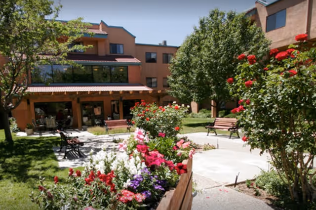 Outdoor garden area at Sandia Vista Senior Living with colorful flowers, green trees, benches, and a multi-story building in the background under a clear blue sky.