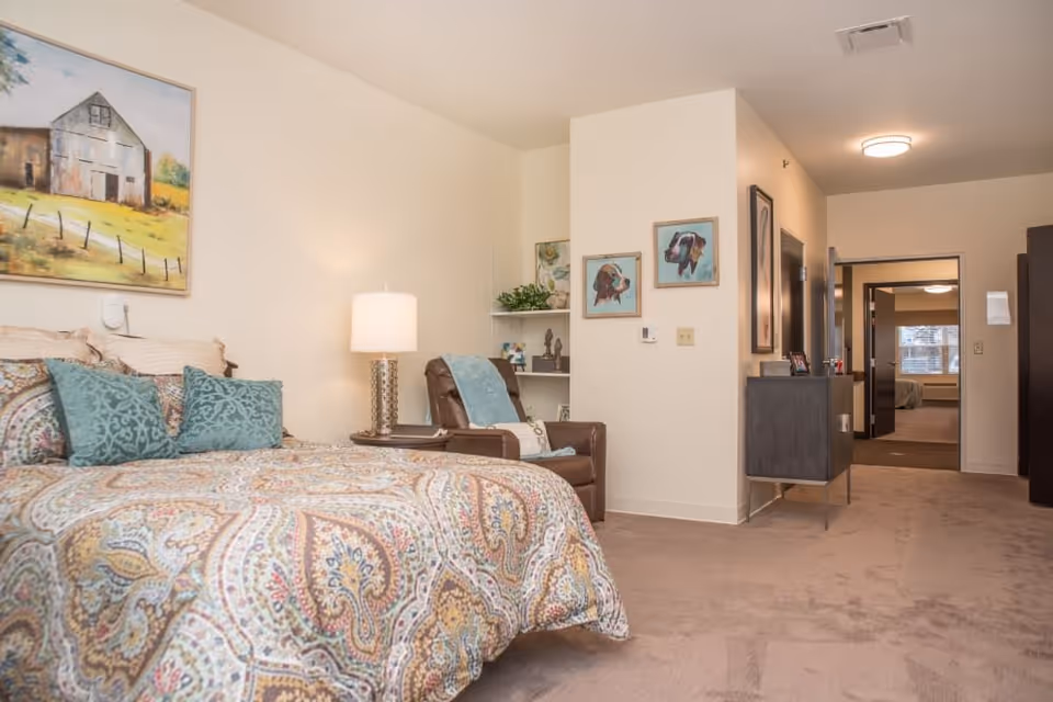 Bright furnished bedroom with a patterned bedspread, armchair and bedside lamp opening to a hallway and adjoining room.