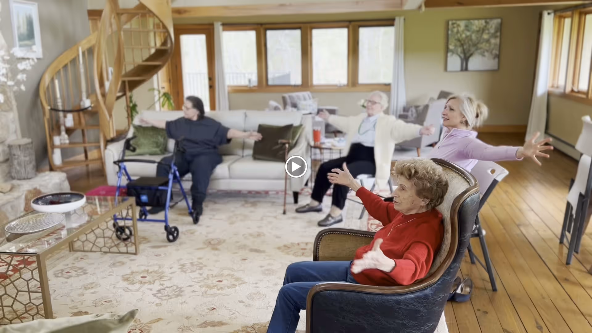 Four elderly women seated in a spacious, well-lit living room with wooden floors and large windows, engaging in a seated stretching exercise with their arms extended. The room features a spiral staircase, a stone fireplace, a glass coffee table, and comfortable seating including a sofa and armchairs.