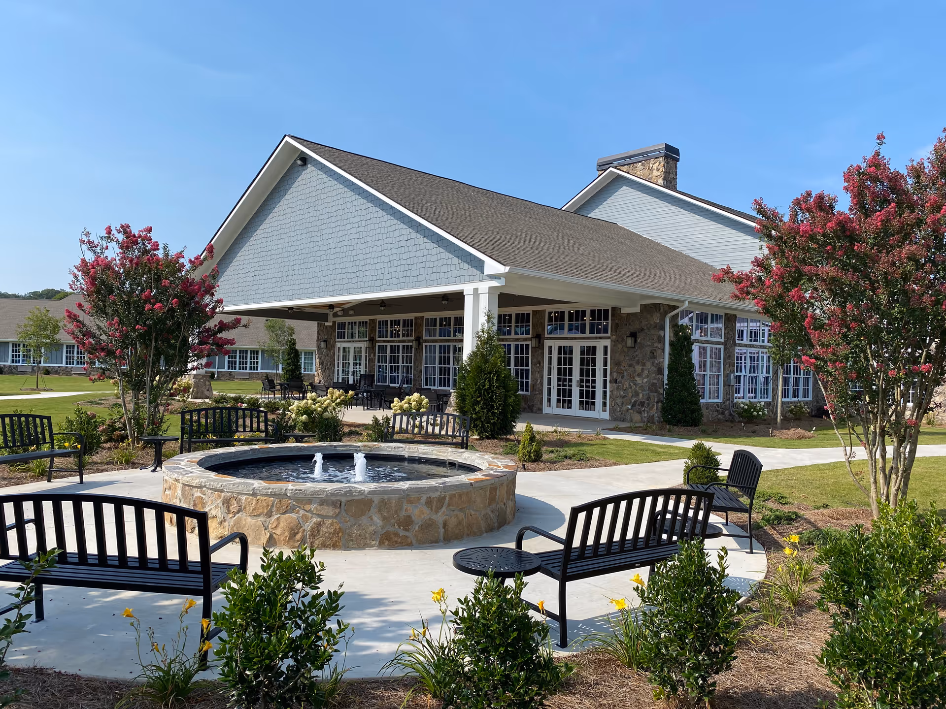 Outdoor seating area with black metal benches arranged around a circular stone water fountain with three small water jets. The area is surrounded by landscaped greenery, flowering bushes, and trees with pink blossoms. In the background, there is a large building with stone and light blue siding, multiple windows, and a covered patio area with tables and chairs under a clear blue sky.