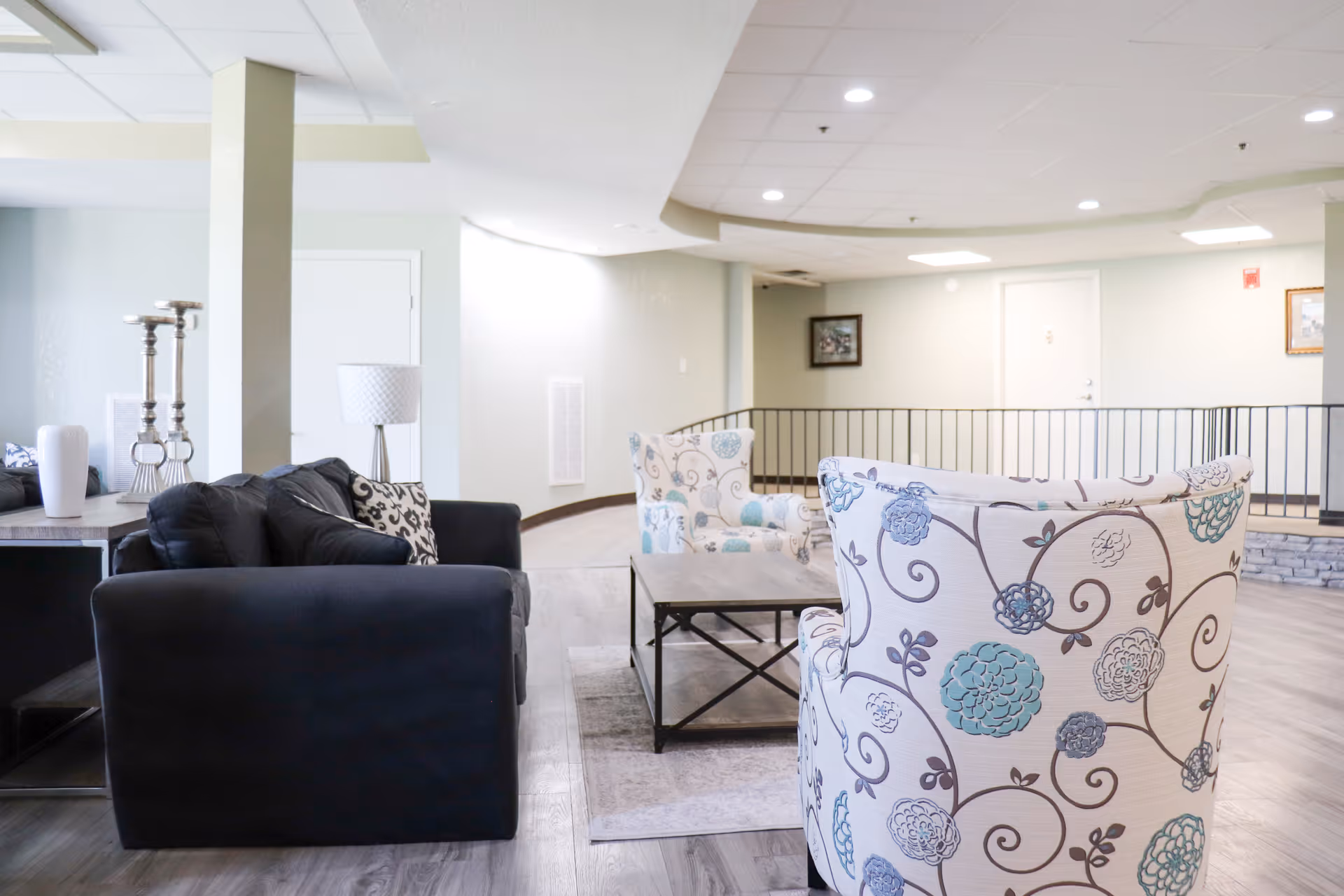 Bright common seating area with patterned armchairs, a dark sofa, coffee table, and decorative lamps.