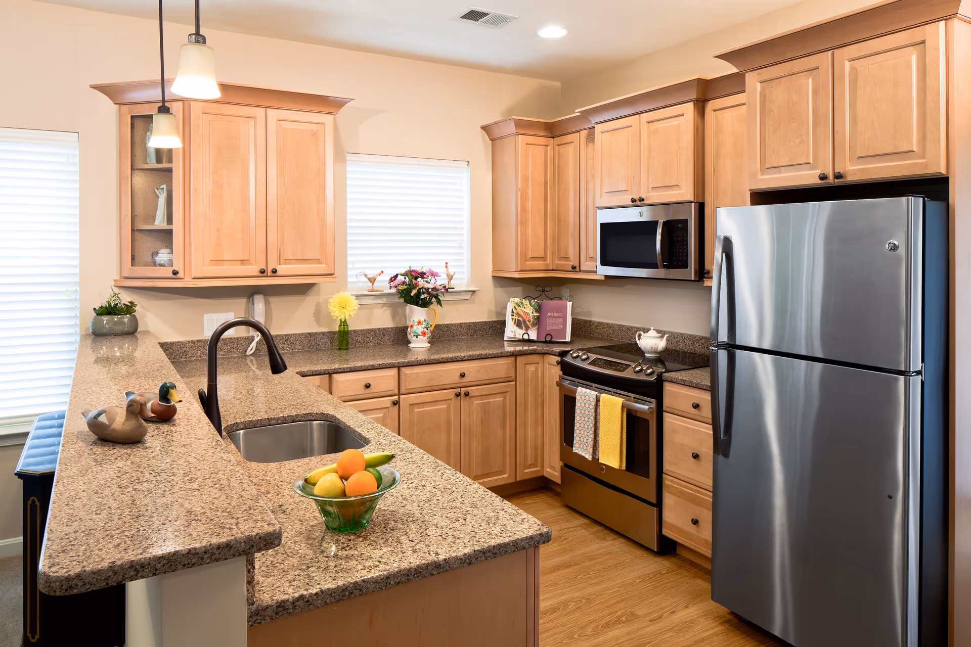 Bright modern kitchen with granite countertops, light wood cabinets, stainless steel refrigerator and stove, and a sink on a peninsula with a bowl of fruit.