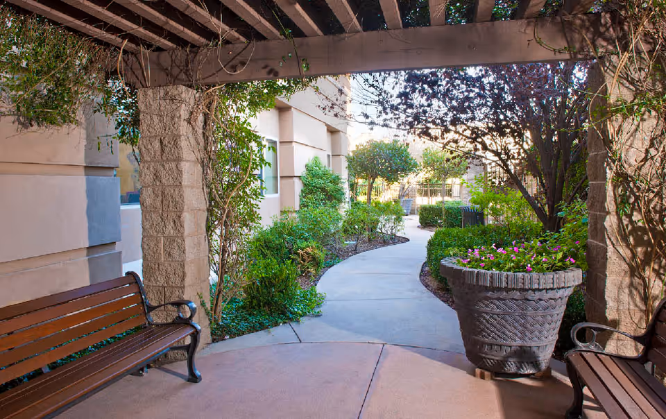 Covered courtyard walkway with benches, a large planter of flowers, and a landscaped path lined with shrubs and trees.