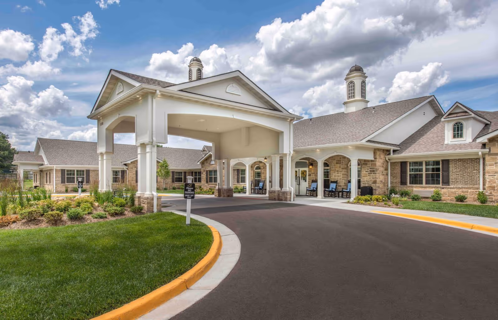 Exterior view of Benton House of Olathe showing a covered entrance with white columns, a curved driveway, manicured green lawn, and a partly cloudy sky.
