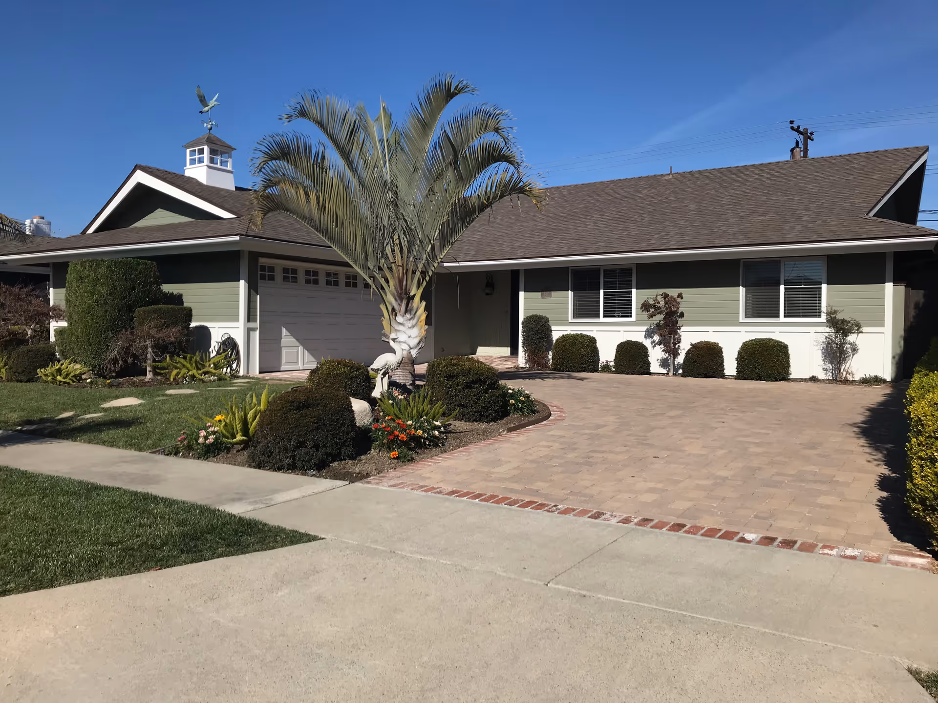 Front exterior of a single-story green house with a garage, paved driveway, palm tree, and landscaped yard under a clear blue sky.