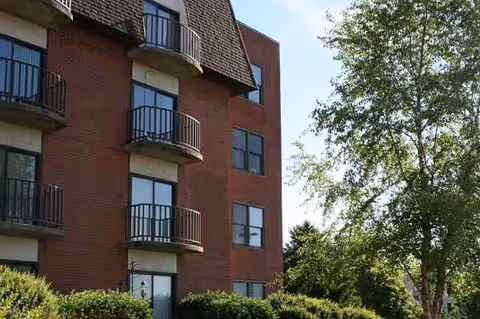 Brick senior living building with multiple balconies, shrubs, and a tree-lined foreground.