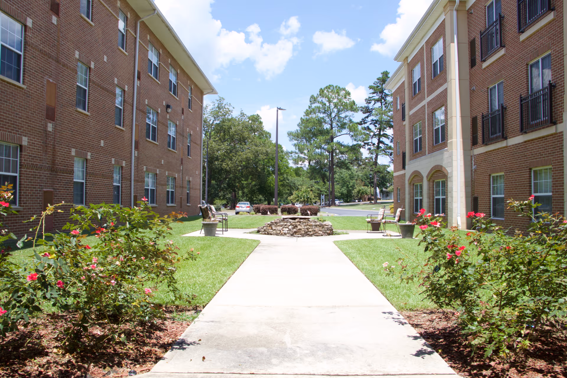 Outdoor courtyard area between two brick buildings with a concrete walkway, green grass, flowering bushes, benches, and a stone fire pit under a partly cloudy blue sky.