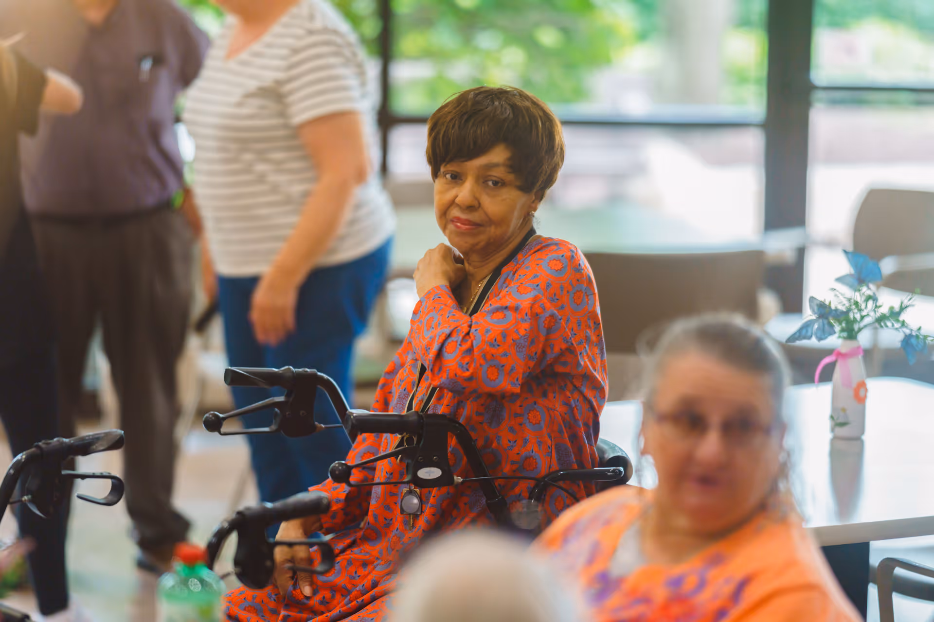 An older woman in a patterned dress sits with a walker in a bright communal dining area while other seniors converse nearby.
