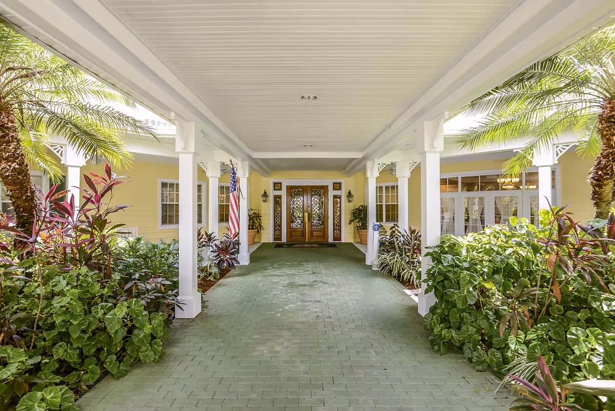 Covered entrance walkway to a building with white pillars and a green tiled floor, surrounded by lush green plants and palm trees, leading to double wooden doors with glass panels. The building exterior is painted yellow with white trim, and an American flag is displayed near the entrance.