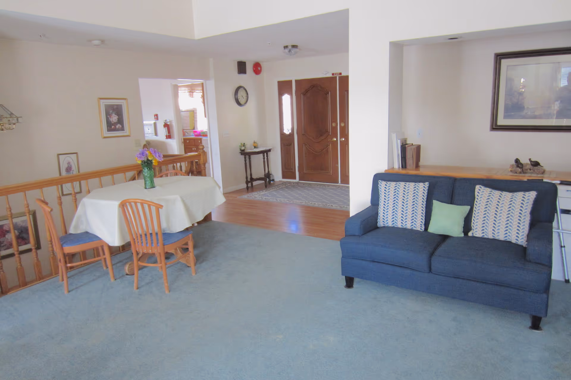 Interior view of a senior living facility showing a small dining area with a table covered by a white tablecloth and four wooden chairs, a blue couch with three decorative pillows, a wooden railing, and a front door area with a small table and a wall clock.