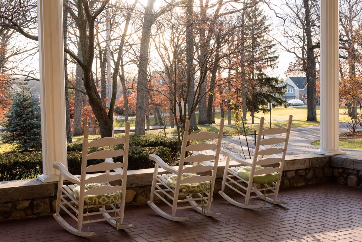 Three white rocking chairs with green cushions sit on a covered porch facing a lawn and trees.