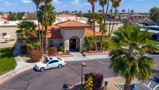 Front entrance of a Mediterranean-style senior living facility with a tiled roof, palm trees, colorful landscaping, and a car in the driveway.