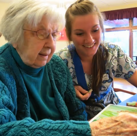 An elderly woman with white hair and glasses wearing a teal sweater is sitting next to a young woman with long brown hair in a patterned shirt. They are both looking at a board game or activity on the table in front of them, smiling and engaged.