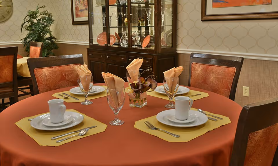 A round dining table set for four with orange tablecloth and yellow placemats, each place setting includes a white cup, saucer, plate, and silverware. There are folded peach-colored napkins in water glasses and a small floral centerpiece. In the background, there is a dark wooden china cabinet with glassware and decorative plates, a potted plant, and framed artwork on the wall.
