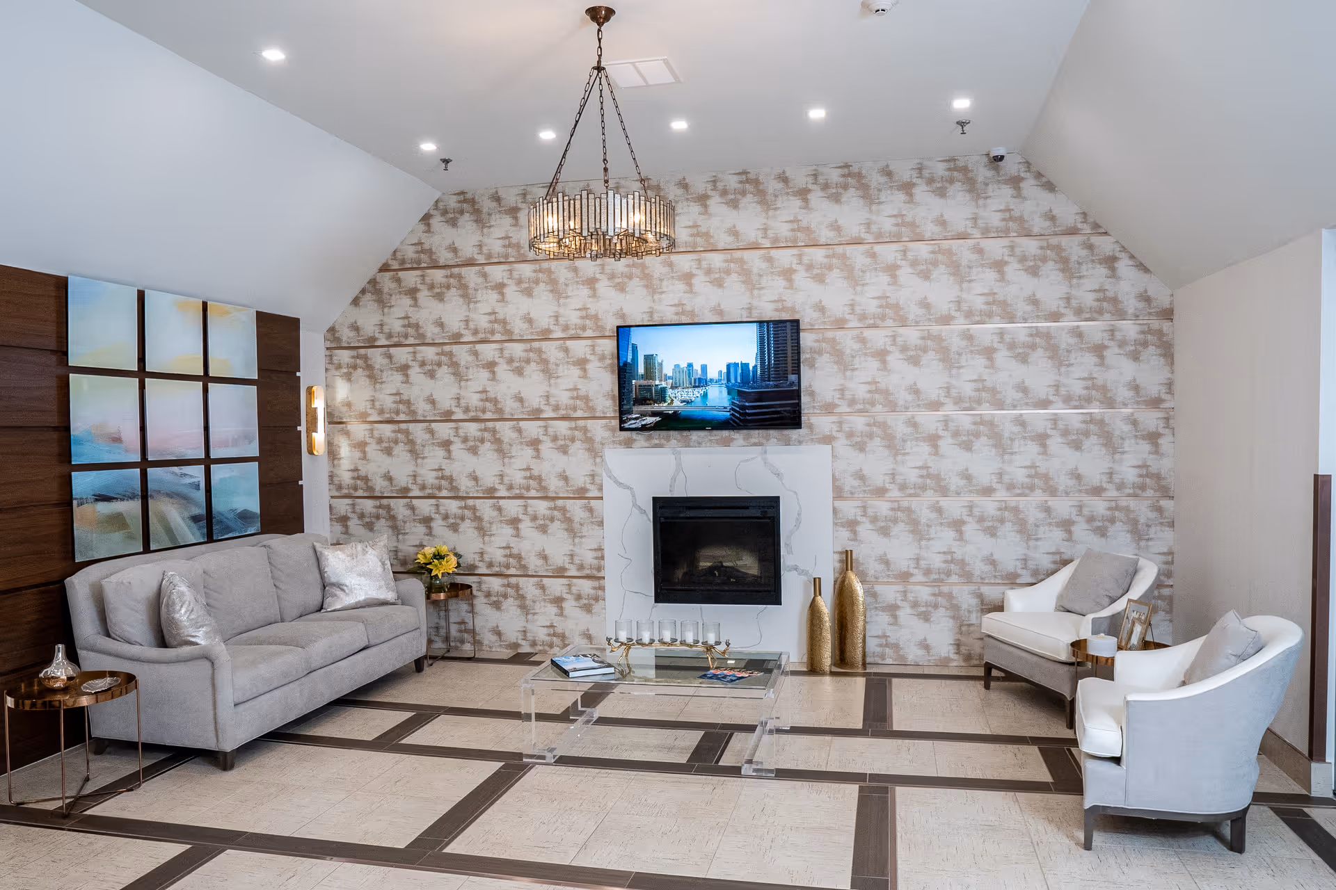A modern living room with a gray sofa, two white armchairs, a glass coffee table, and a fireplace with a mounted flat-screen TV above it. The walls have patterned wallpaper and a wooden panel with abstract artwork. A chandelier hangs from the ceiling.