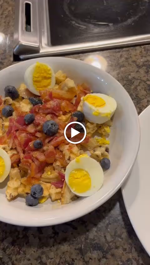 Bowl of scrambled eggs topped with bacon, blueberries and halved hard-boiled eggs on a kitchen counter next to a stovetop.