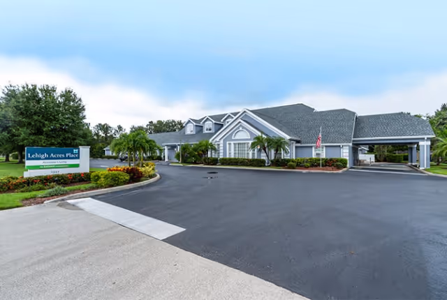 Exterior view of Lehigh Acres Assisted Living facility showing a single-story building with a gray roof, white trim, and an American flag near the entrance. The driveway is freshly paved and there are landscaped bushes and palm trees around the building. A green and white sign with the facility name is visible on the left side near the road.