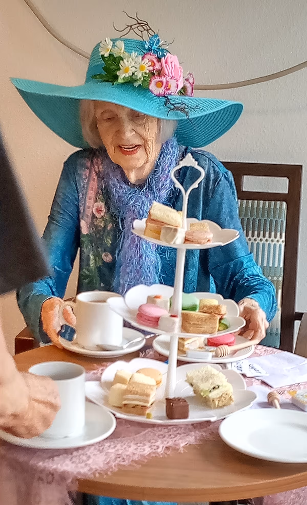 An elderly woman wearing a large turquoise hat decorated with flowers and a blue scarf is sitting at a table with a tiered tray of assorted small sandwiches and pastries. She is holding a white cup and saucer, and there are additional cups and plates on the table covered with a pink lace tablecloth.
