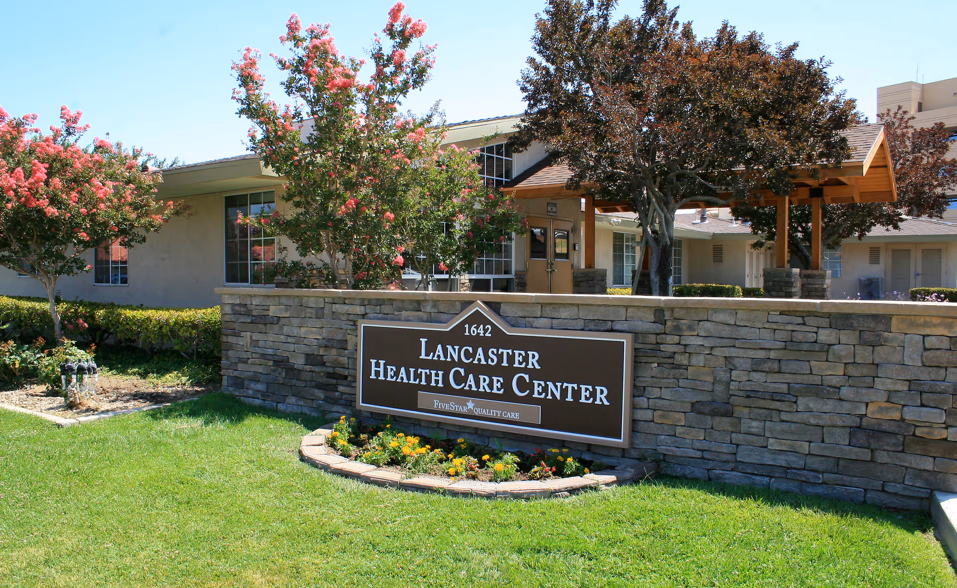 Exterior view of Lancaster Health Care Center showing a stone wall with a sign displaying the facility's name and address, surrounded by green grass, flowering trees, and a covered entrance.