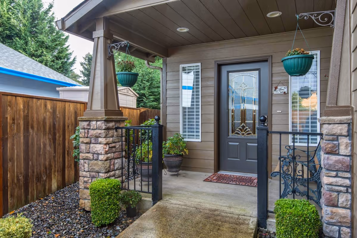 Front porch entrance of a house with a decorative glass door, two narrow windows with white blinds on either side, hanging green flower pots, stone pillars, black metal railing, potted plants, and a wooden fence surrounding the area.