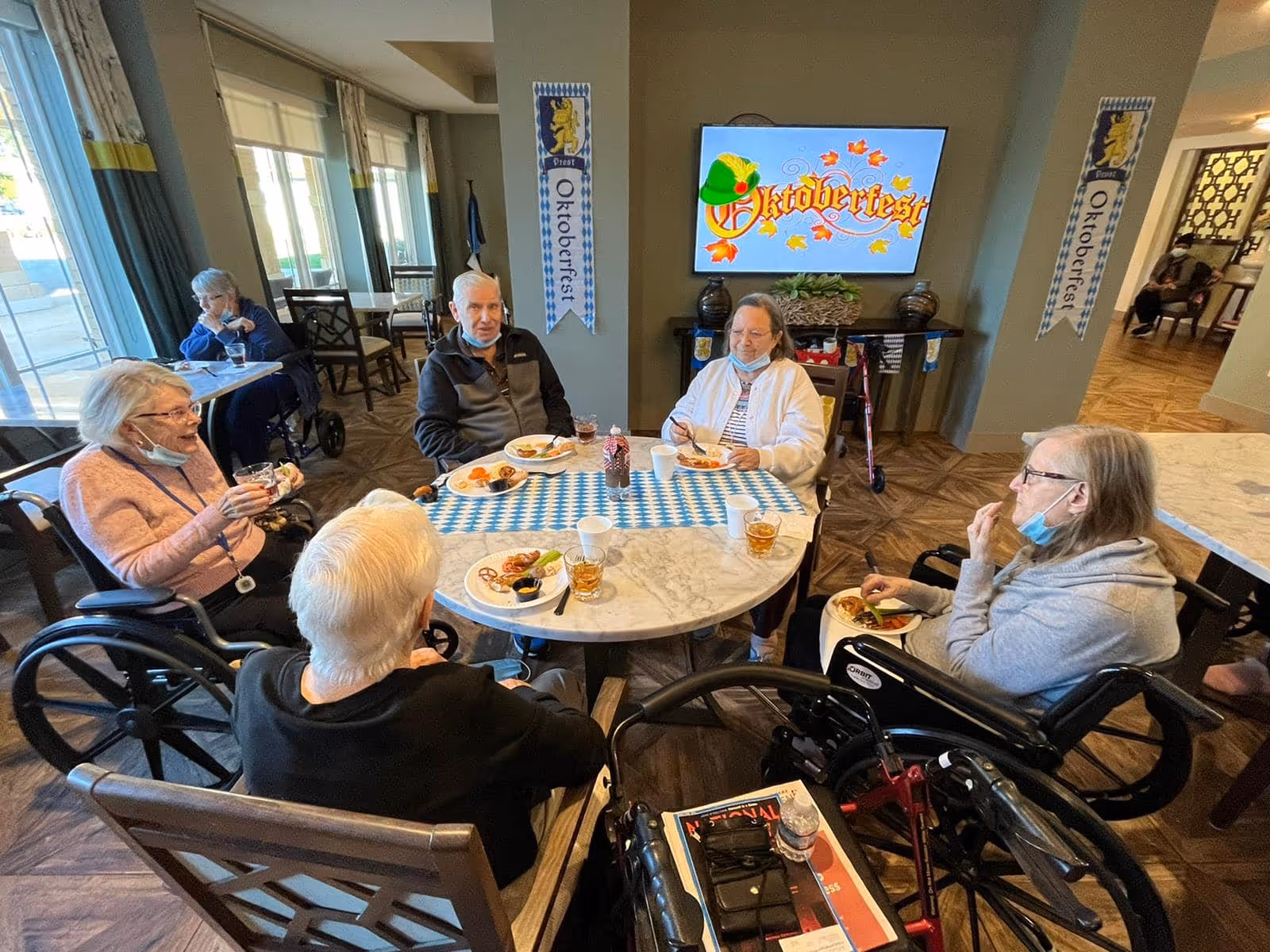 Several elderly residents sit around a table eating and socializing in a decorated common dining area with an 'Oktoberfest' sign on a wall-mounted TV.