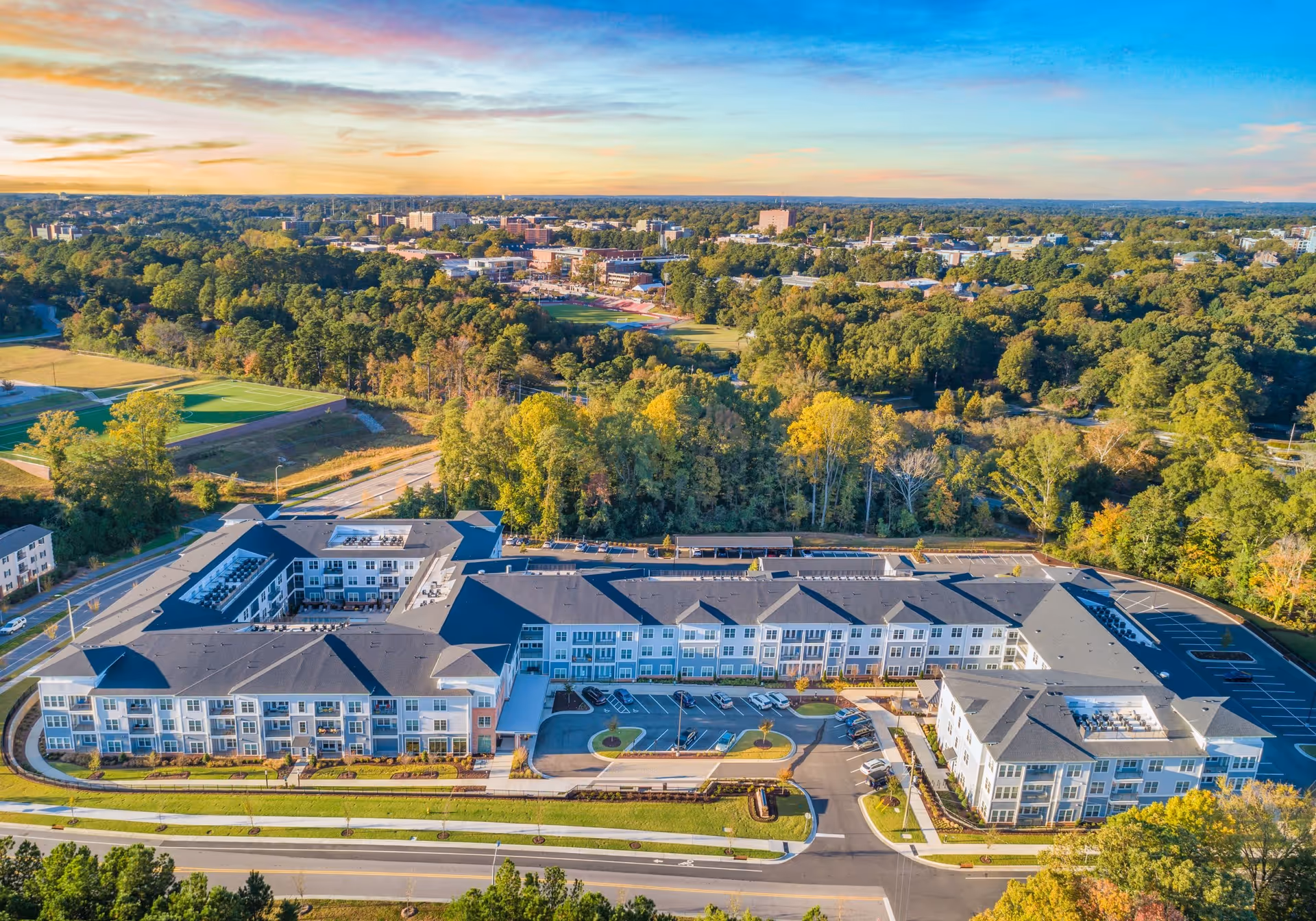 Aerial view of Overture Centennial, a large senior living facility surrounded by trees and greenery, with a parking lot and roads in the foreground and a cityscape in the background under a partly cloudy sky during sunset.