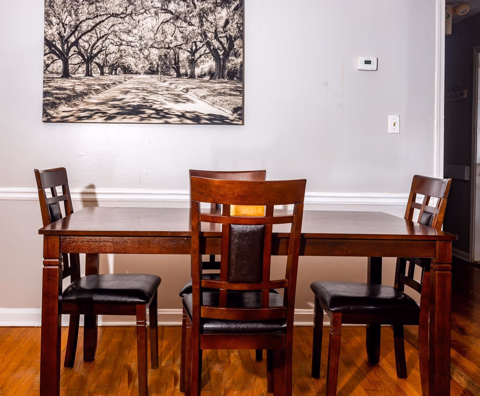A wooden dining table with four matching chairs featuring black cushioned seats, set on a hardwood floor. On the wall behind the table is a large black and white photograph of a tree-lined path. The wall is painted light gray with white trim, and there is a thermostat and light switch visible on the right side.