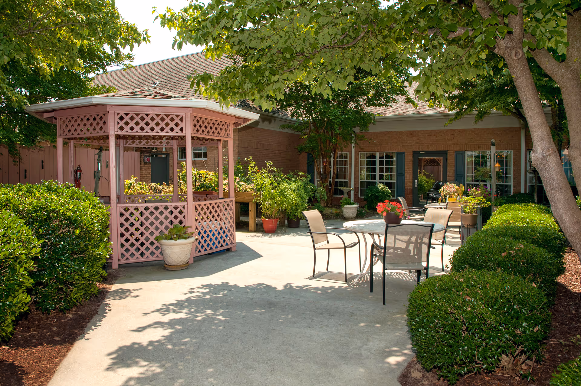 Outdoor patio area at a senior living facility with a pink gazebo, several potted plants, round tables with chairs, and green bushes and trees providing shade.