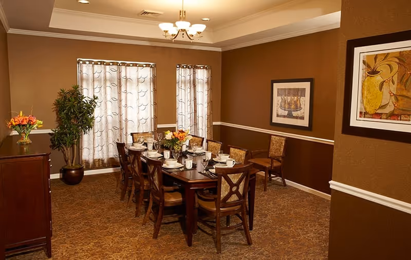 A dining room with a long wooden table set for a meal, surrounded by ten wooden chairs with cushioned seats. The room has brown walls with white trim, two framed paintings on the walls, a chandelier hanging from the ceiling, and two windows with patterned curtains. There is a potted plant and a sideboard with a vase of flowers on the left side.