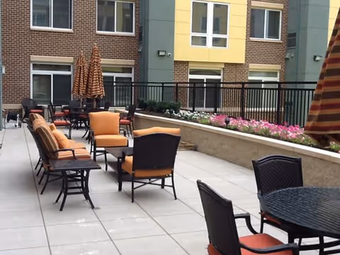 Outdoor patio area with multiple tables and chairs featuring orange cushions, surrounded by a low wall with pink flowers and a multi-story building with brick and yellow siding in the background.