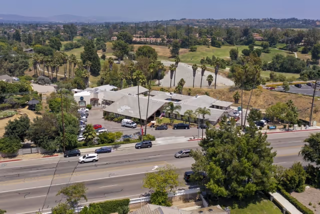 Aerial view of St. Elizabeth Healthcare & Rehab facility surrounded by trees and greenery, with a parking lot filled with cars and a main road with vehicles in the foreground. The building has a light-colored roof and is situated near a golf course or open green area.