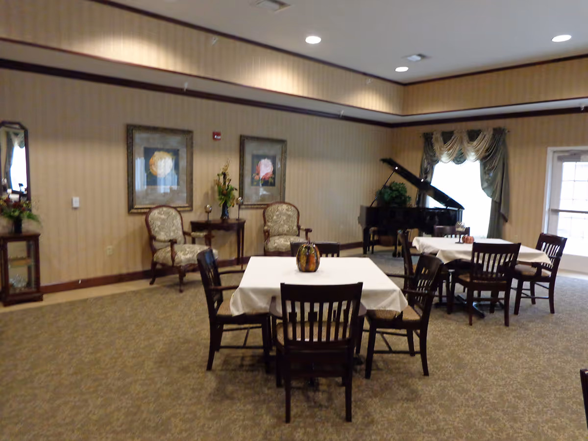 A common area with two tables covered with white tablecloths, each surrounded by four wooden chairs. In the background, there are two upholstered armchairs with a small table between them, decorative framed flower pictures on the wall, a grand piano near a window with draped curtains, and a door leading outside.