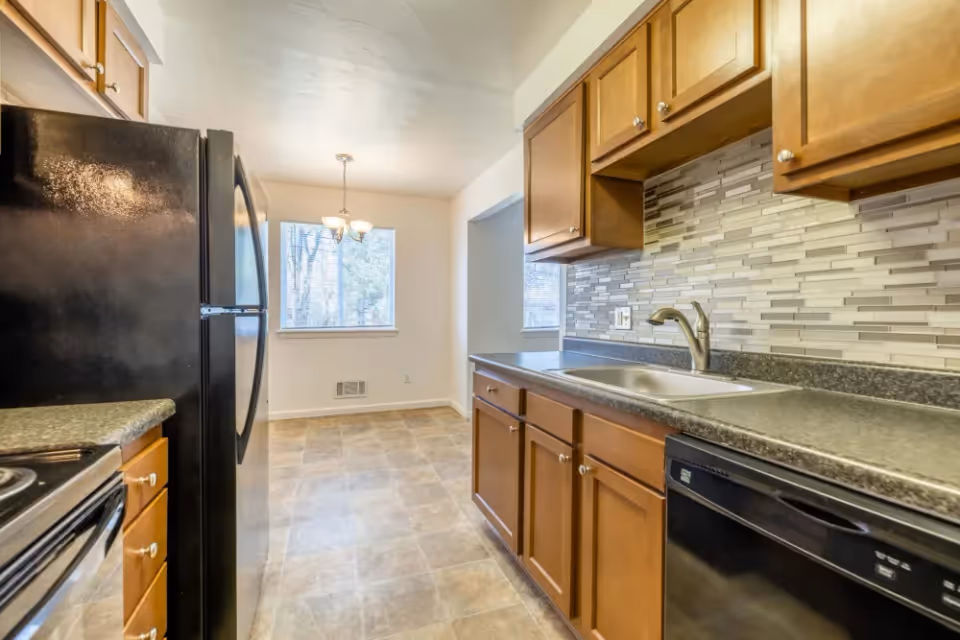 A kitchen with wooden cabinets, a black refrigerator, a black dishwasher, a sink with a faucet, and a tiled backsplash. The kitchen opens into a dining area with a window and a hanging light fixture.