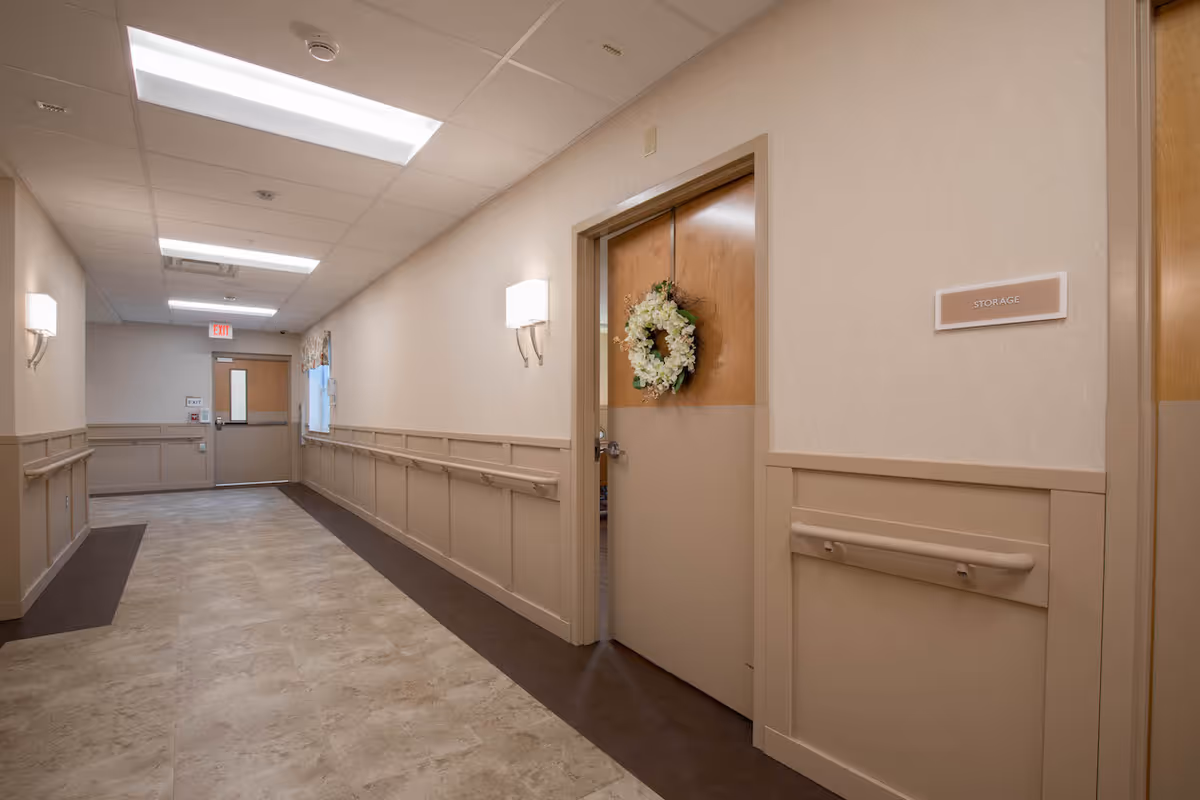 A clean and well-lit hallway in a nursing and rehabilitation center with beige walls, handrails, and a door on the right labeled 'Storage' decorated with a floral wreath. The hallway has a tiled floor and fluorescent ceiling lights, with an exit door visible at the far end.
