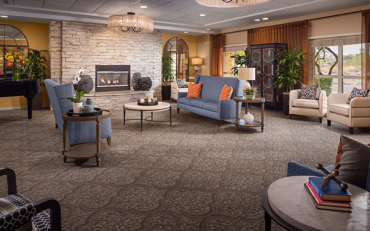 A spacious and well-lit senior living facility lounge area featuring a stone fireplace with a fire burning, surrounded by comfortable seating including blue and beige armchairs and sofas with orange and patterned cushions. The room has large windows with brown curtains, decorative plants, a round coffee table, side tables with lamps, and a patterned carpet. There is also a black grand piano and decorative mirrors on the walls.